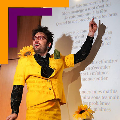 Visuel du spectacle "Le Chantoir Public". Un personnage en tailleur jaune et boa, entouré de tournesols, se trouve devant un tableau avec des paroles de chansons.
