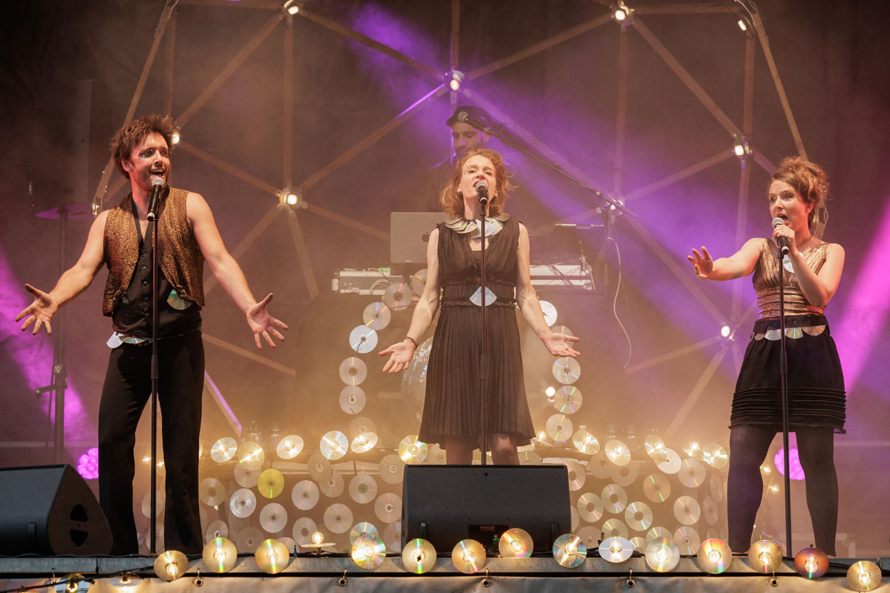 La photo représente 3 chanteurs sur une scène sous une sorte de géode avec beaucoup de lumières, teintes naturelles et dans les tons violine.