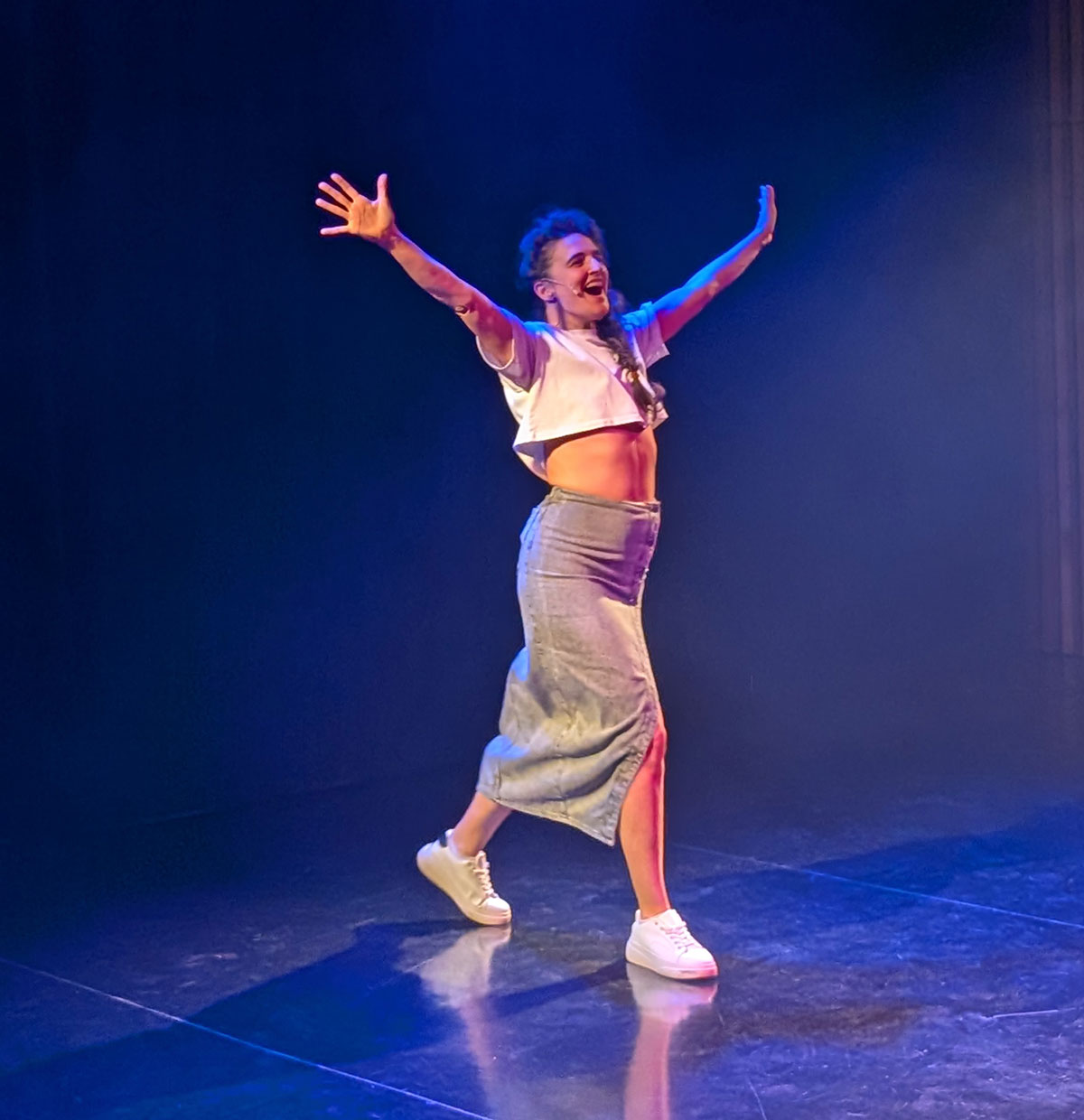 Visuel pour le spectacle Pilule Rouge. On y voit une femme seule sur la scène, dans la lumière bleu, qui semble satisfaite d'une victoire, elle lève les bras en s'exclamant.
