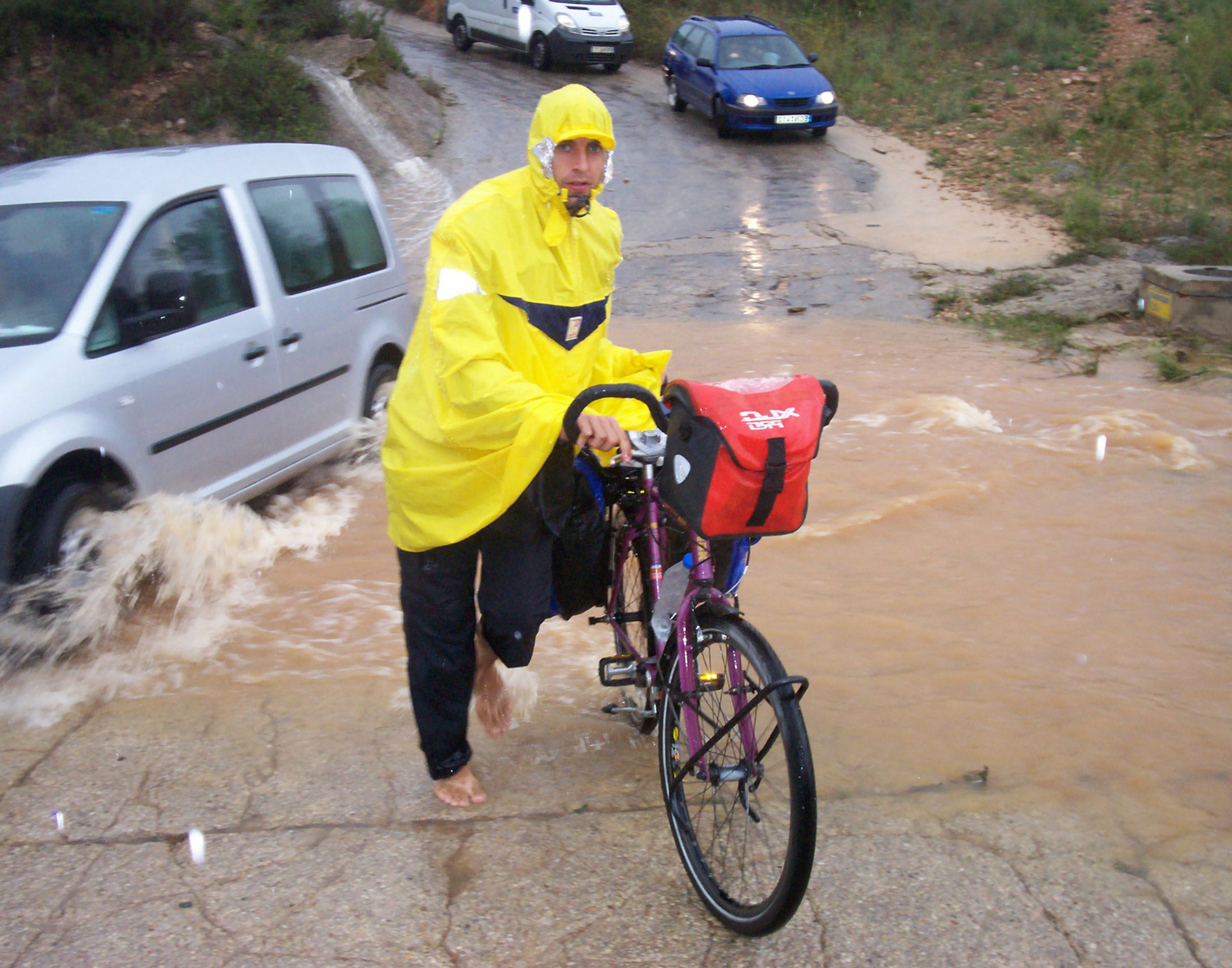 Visuel qui illustre le spectacle "Apartés". On y voit un cycliste en k-way jaune, poussé son vélo, pieds nus sous une pluie battante.