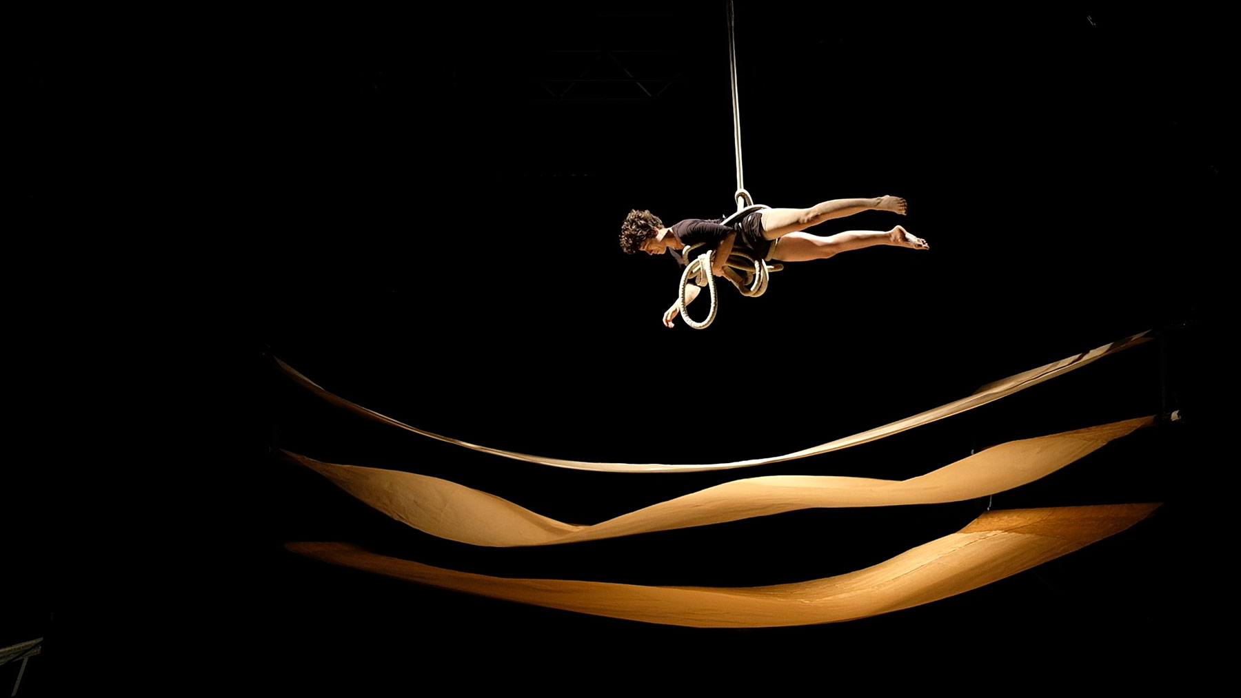 Visuel qui illustre le spectacle "Pli". On y voit une acrobate suspendue au dessus de long rubans de papier qui flottent horizontalement dans une belle lumière dorée.