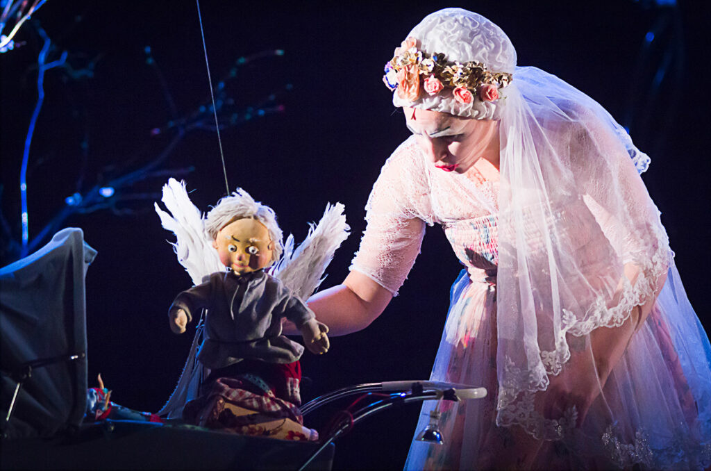 Le visuel illustre le spectacle "Les enfants c'est moi". Il montre une femme clown, vêtue en mariée, penchée sur un berceau avec à la main une marionnette d'ange.