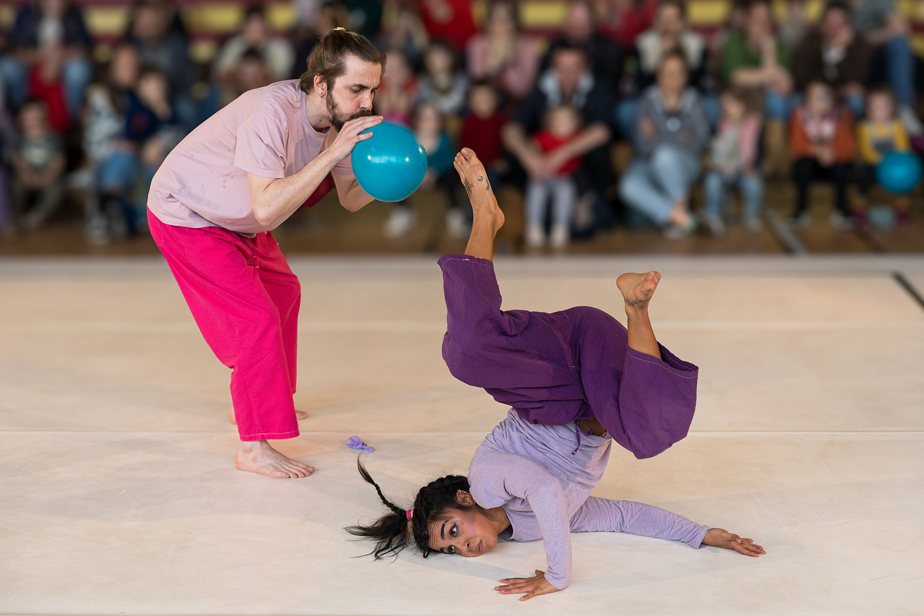 Ce visuel du spectacle Baoum présente sur la scène un artiste qui fait des bruits de bouche avec un ballon de baudruche bleu, devant une artiste qui réalise au sol une acrobatie de hip hop. Le public est assis tout autour de l'espace scénique.