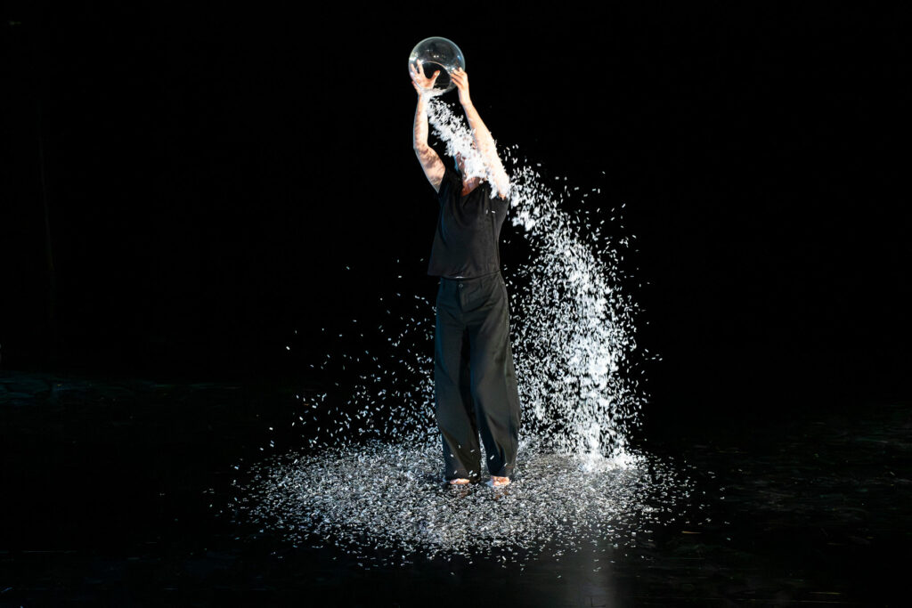 La photographie illustre le spectacle "Sous mes nuits blanches". Elle montre un personnage qui se trouve sous une pluie de confettis de papier qui tombent d'un bocal. C'est la métaphore de la glace qui fond et ruisselle comme de l'eau en cascade.