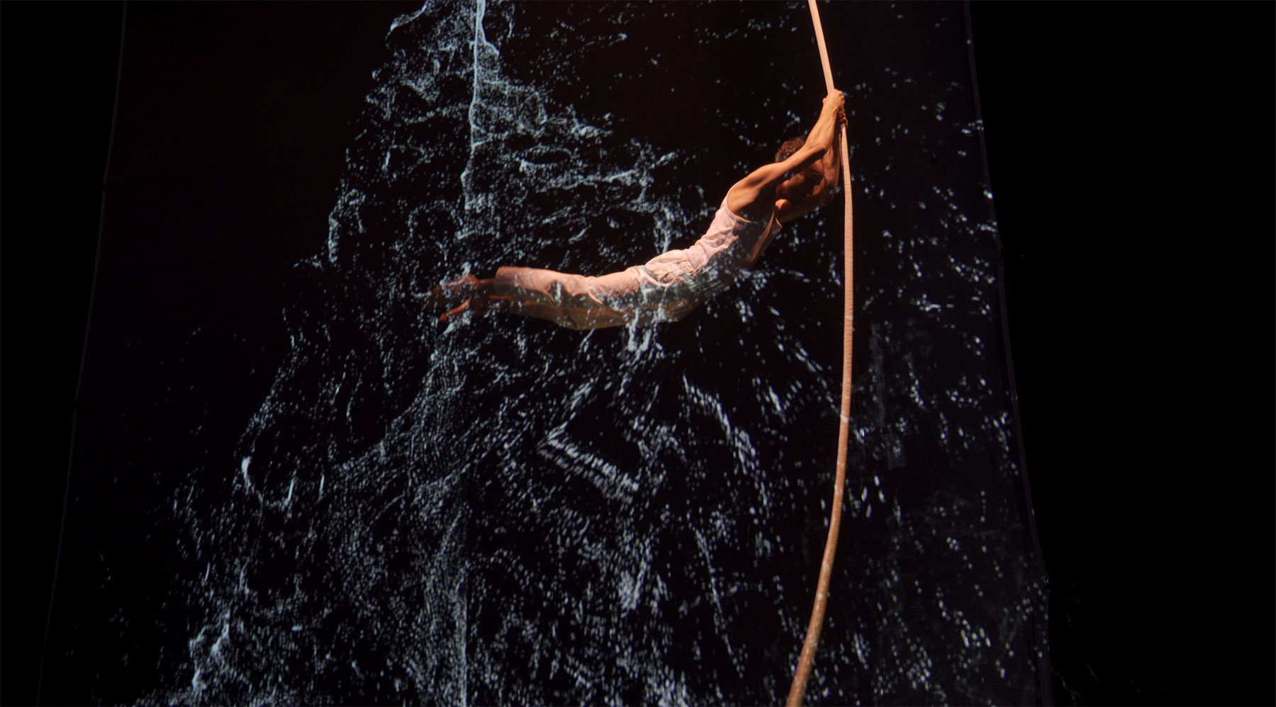 Ce visuel montre un acrobate vêtut de blanc se balançant à l'horizontale sur une corde lisse. Une projection vidéo donne l'impression qu'il traverse une chute d'eau.