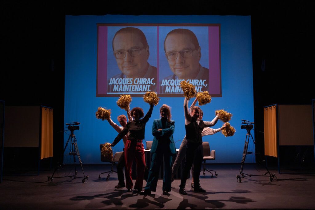 On voit des personnages en costume sombre qui agitent des pompons de pompom girls dorés. Ils se trouvent dans une salle obscure, devant un écran avec des affiches de campagne de Jacques Chirac