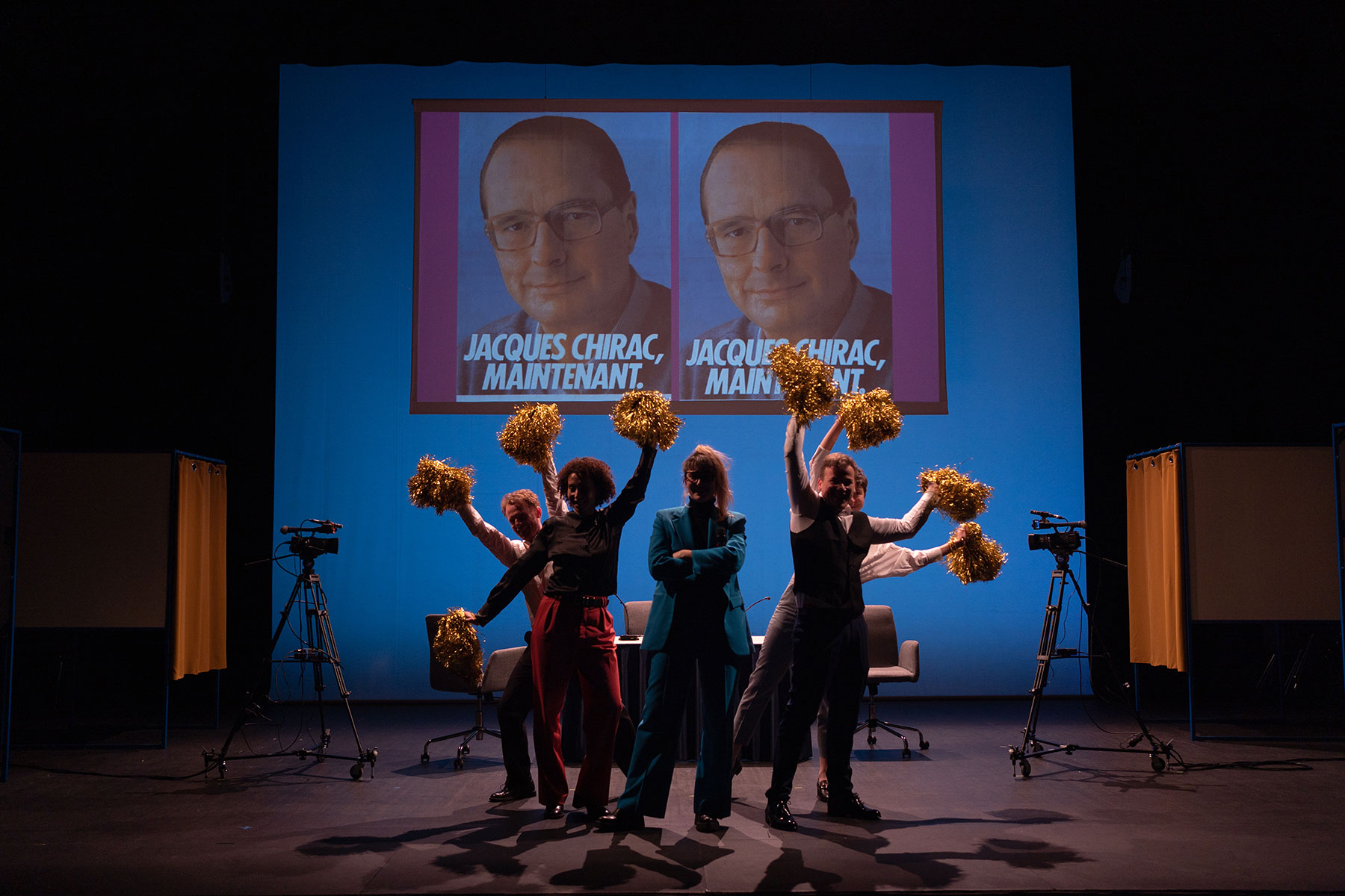 On voit des personnages en costume sombre qui agitent des pompons de pompom girls dorés. Ils se trouvent dans une salle obscure, devant un écran avec des affiches de campagne de Jacques Chirac
